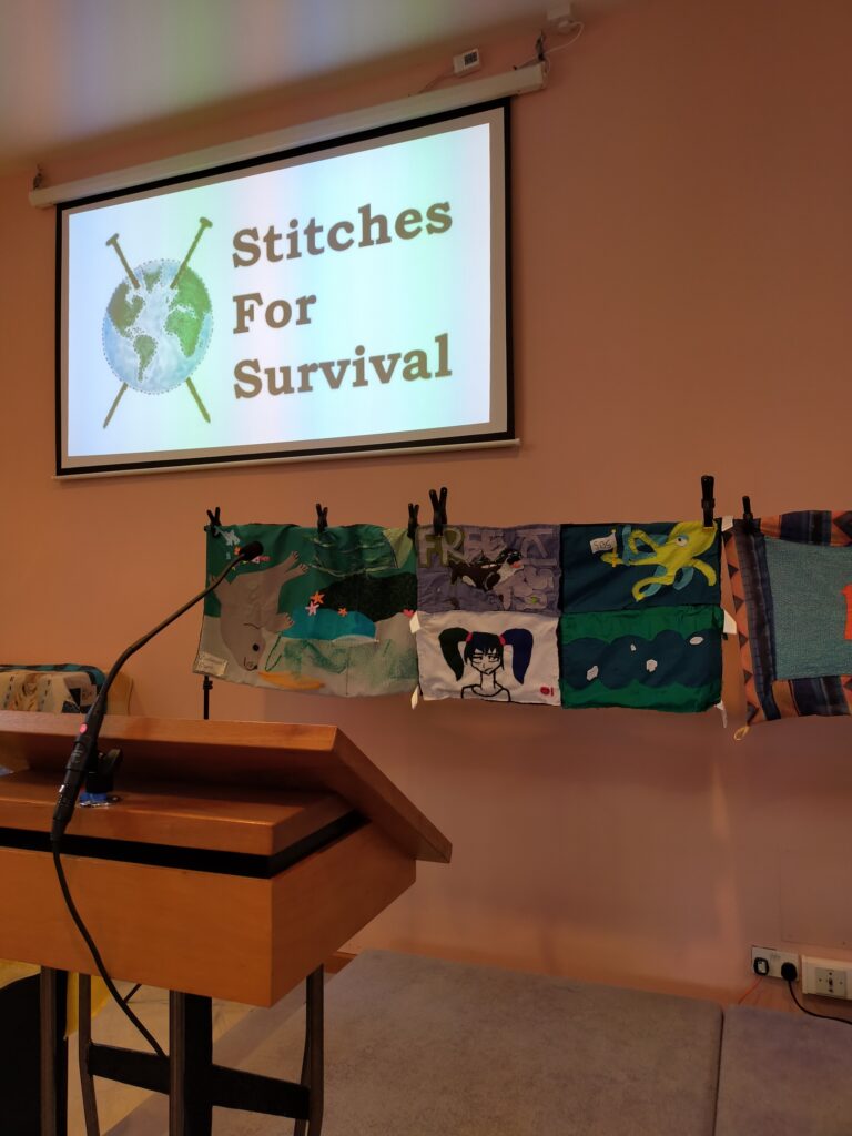 A closeup of the part of the scarf that features SOLE students'  work. The panel is divided in 4. One panel has a killer whale that says "Free Kiska," one has an octopus waving a white flag, one shows an anime-style girl, and one shows a river with ice breaking up in it. Above the scarf is a powerpoint that says "Stitches For Survival" with a pair of knitting needles through an earth.